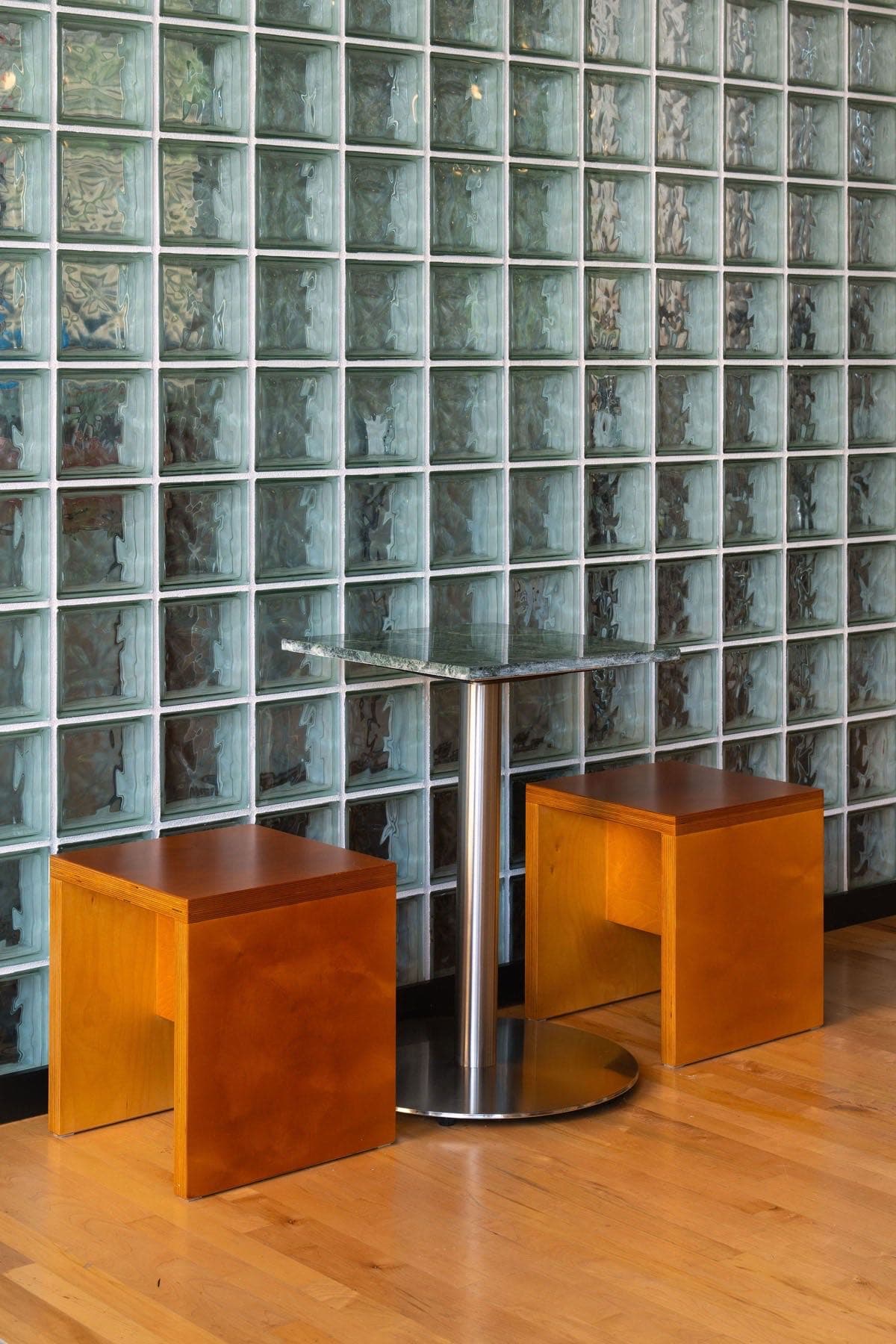 A glass-block wall behind a marble-topped table and two wooden cube stools on the maple floor at Locke Coffee in Newport, KY