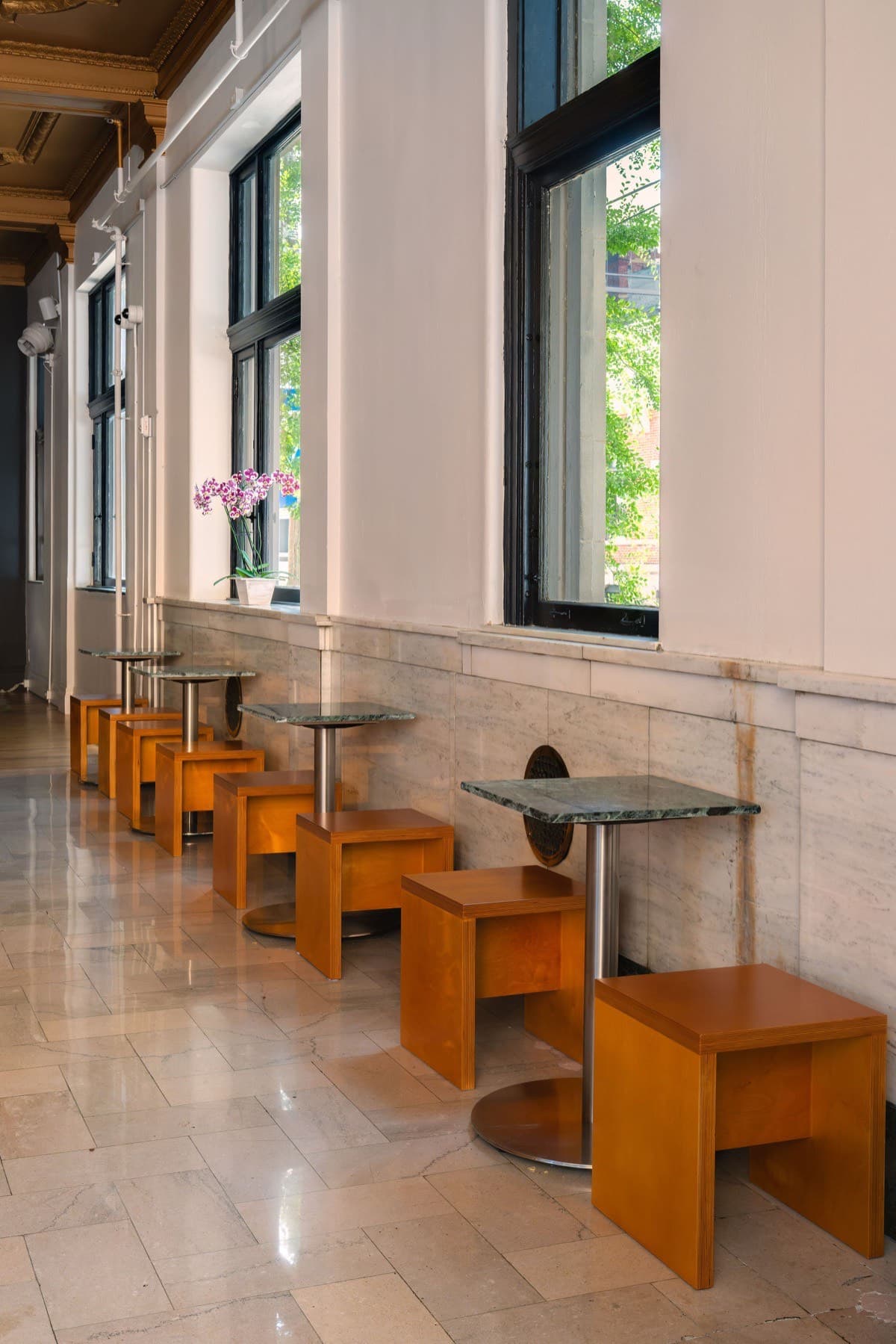 Small square marble-topped tables on chrome pedestals lined up along the window wall at Locke Coffee, with an orchid on the sill and marble tile floor, in Newport, KY near Cincinnati